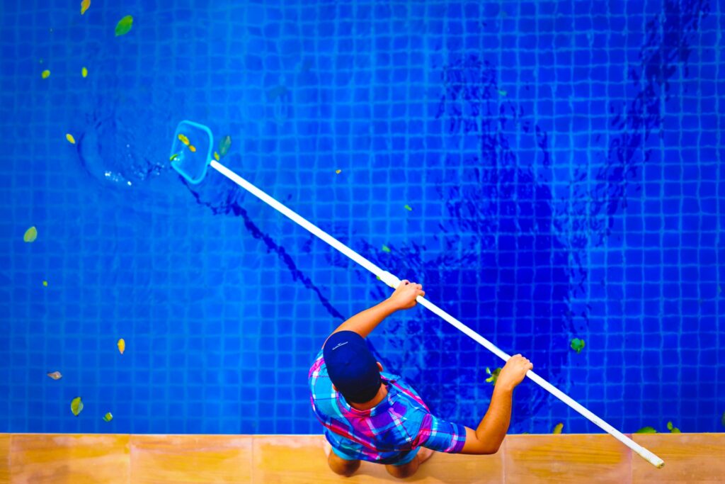 Pool technician skimming leaves from a clear blue swimming pool with a net.