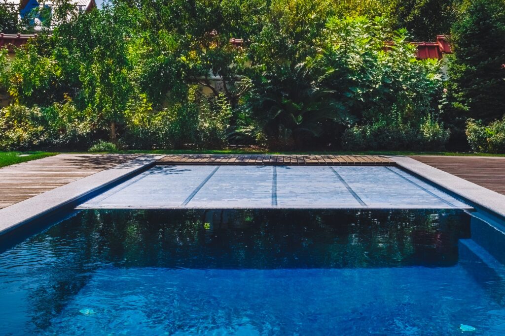 Backyard swimming pool with a partially closed automatic pool cover surrounded by greenery.
