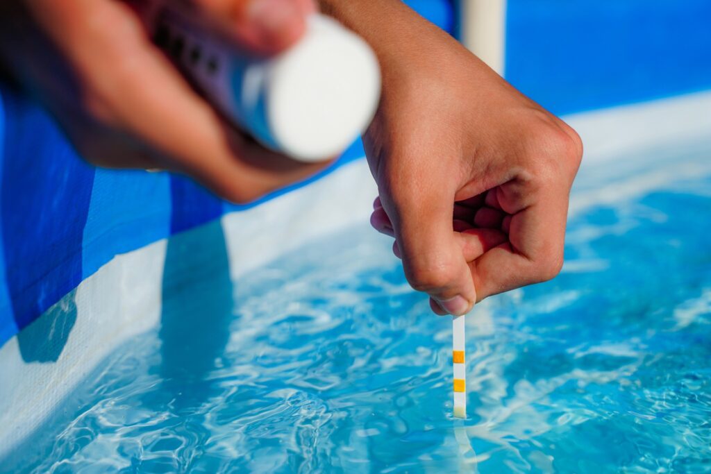 Hand dipping a water test strip into a swimming pool to check chemical balance.