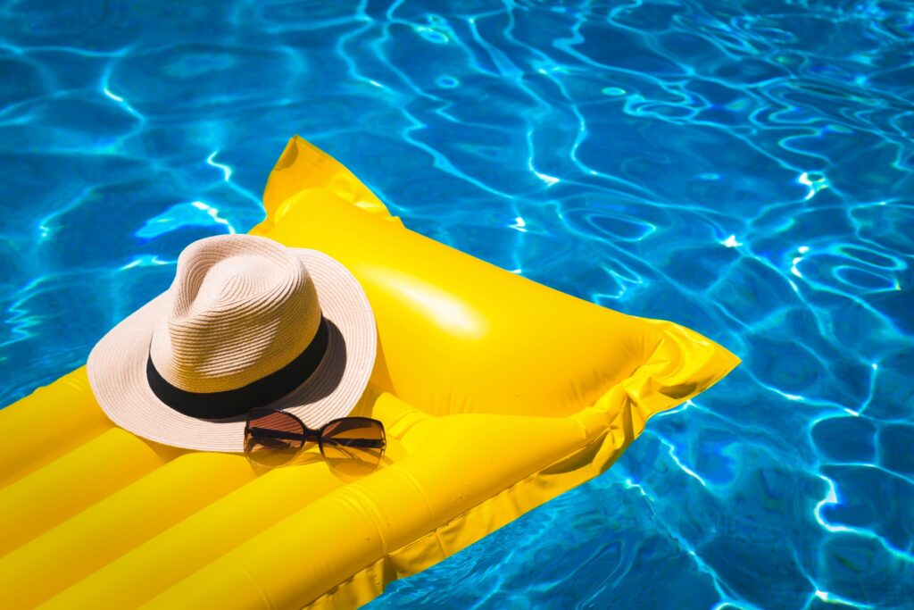 Straw hat and sunglasses resting on a yellow pool float in a bright blue swimming pool.
