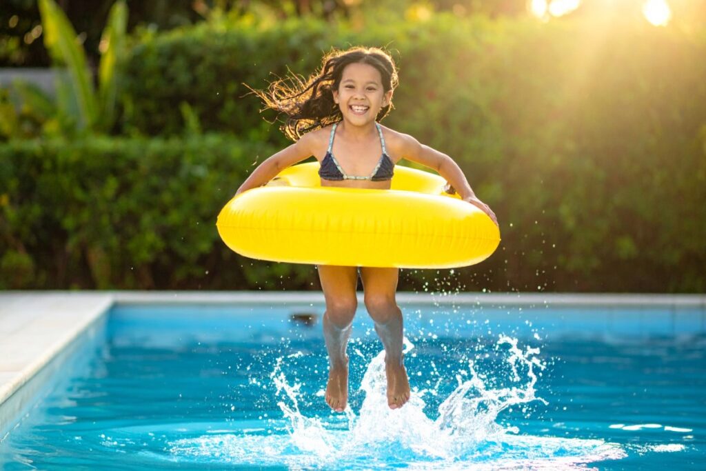 Smiling child jumping into a backyard pool with a yellow float ring on a sunny day.