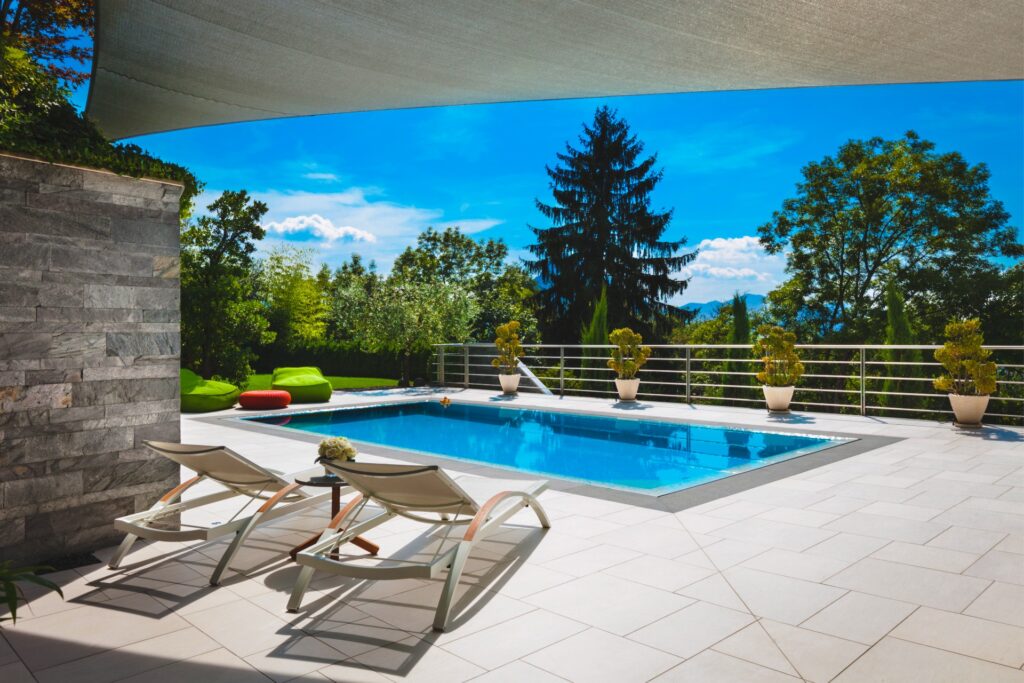A modern outdoor living area with a swimming pool, lounge chairs, shade sail, and landscaped greenery under a bright blue sky.