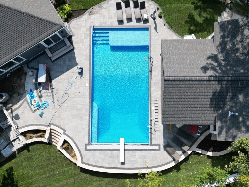 Aerial view of a modern rectangular inground swimming pool surrounded by a light gray paver patio, lounge chairs, and landscaped backyard.