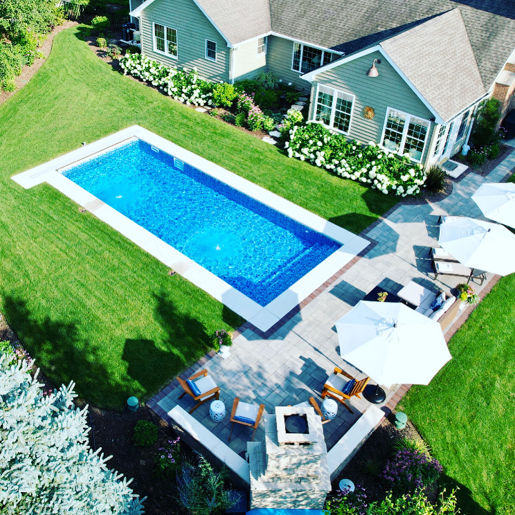 Aerial view of a custom rectangular inground swimming pool with a paver patio, outdoor seating, umbrellas, and landscaped backyard.