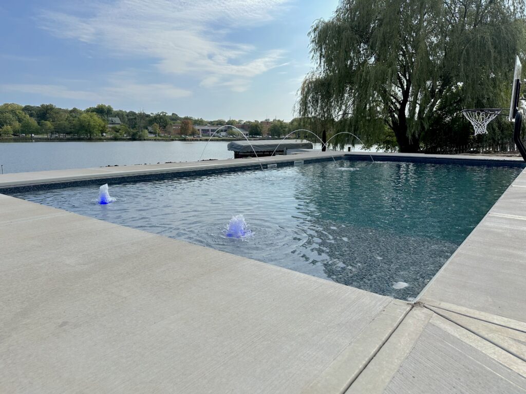 Aerial view of a residential inground swimming pool with a surrounding patio and open green lawn in a landscaped backyard.
