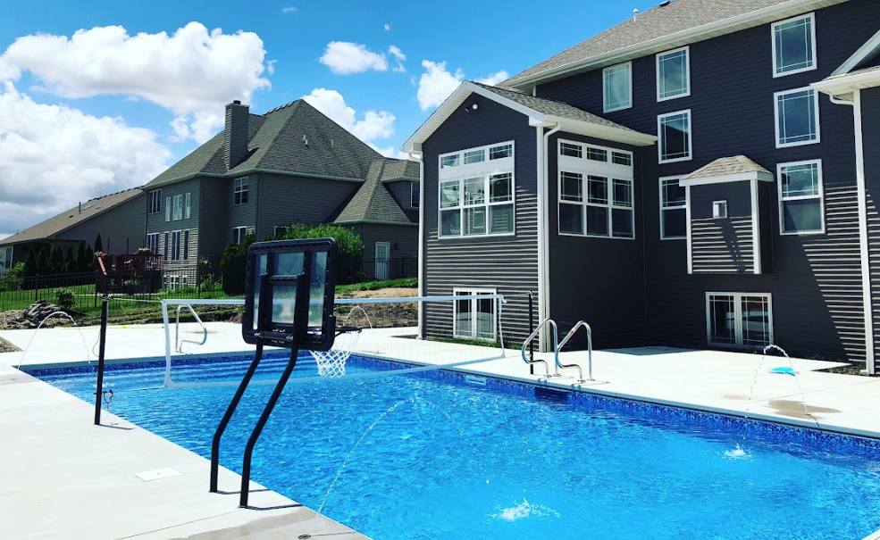 Residential inground swimming pool with a concrete deck, poolside basketball hoop, and multi-story home in the background on a sunny day.