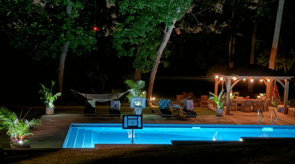 Nighttime view of an illuminated inground swimming pool with underwater lighting, poolside basketball hoop, and outdoor seating area surrounded by trees.