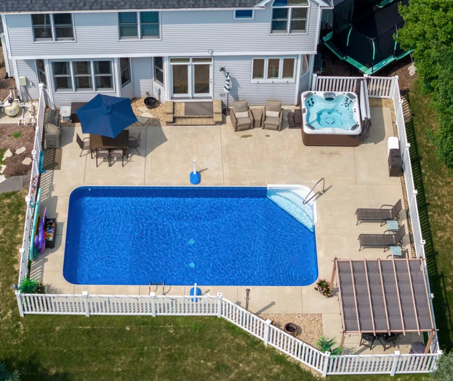 Aerial view of a residential inground swimming pool with a concrete deck, adjacent hot tub, patio seating, and fenced backyard.