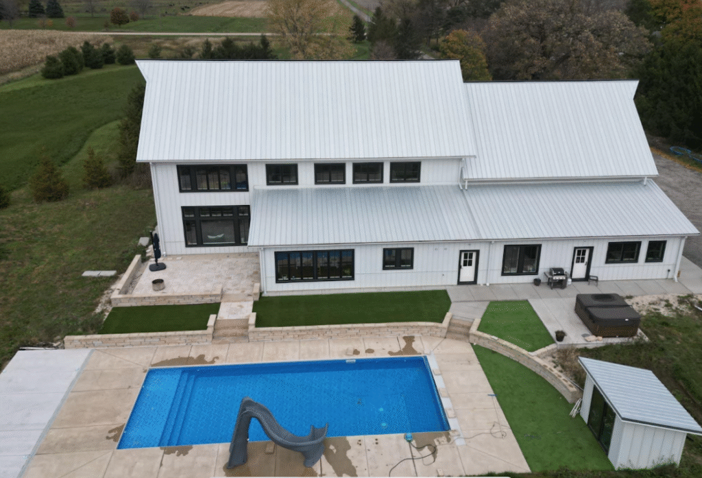 Aerial view of a farmhouse-style home with a rectangular inground swimming pool, concrete deck, slide, and tiered lawn areas.