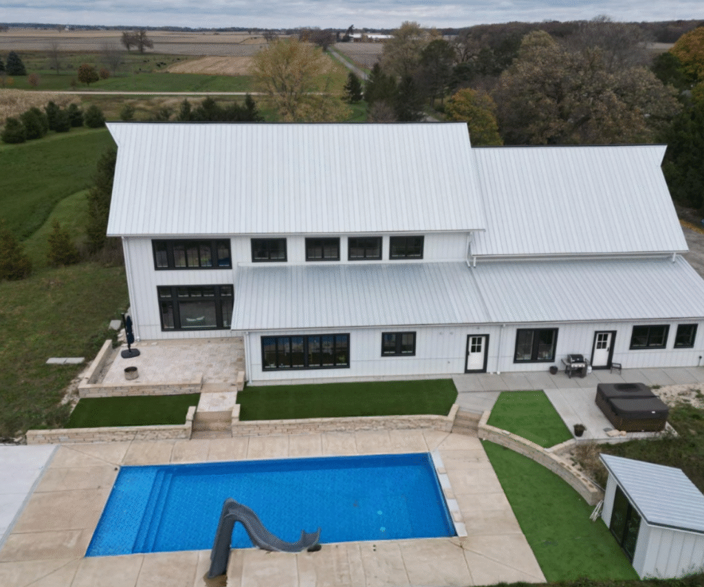 Aerial view of a farmhouse-style home with a rectangular inground swimming pool, concrete pool deck, and surrounding landscaped yard.